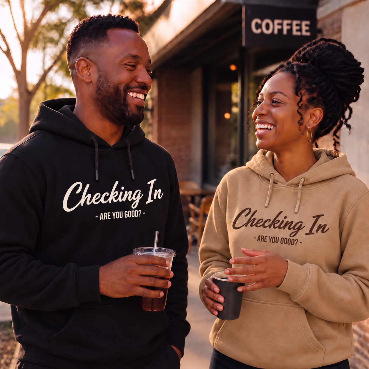 A Black man and woman wearing black and tan “Checking In — Are You Good?” hoodies, laughing together outside a coffee shop.